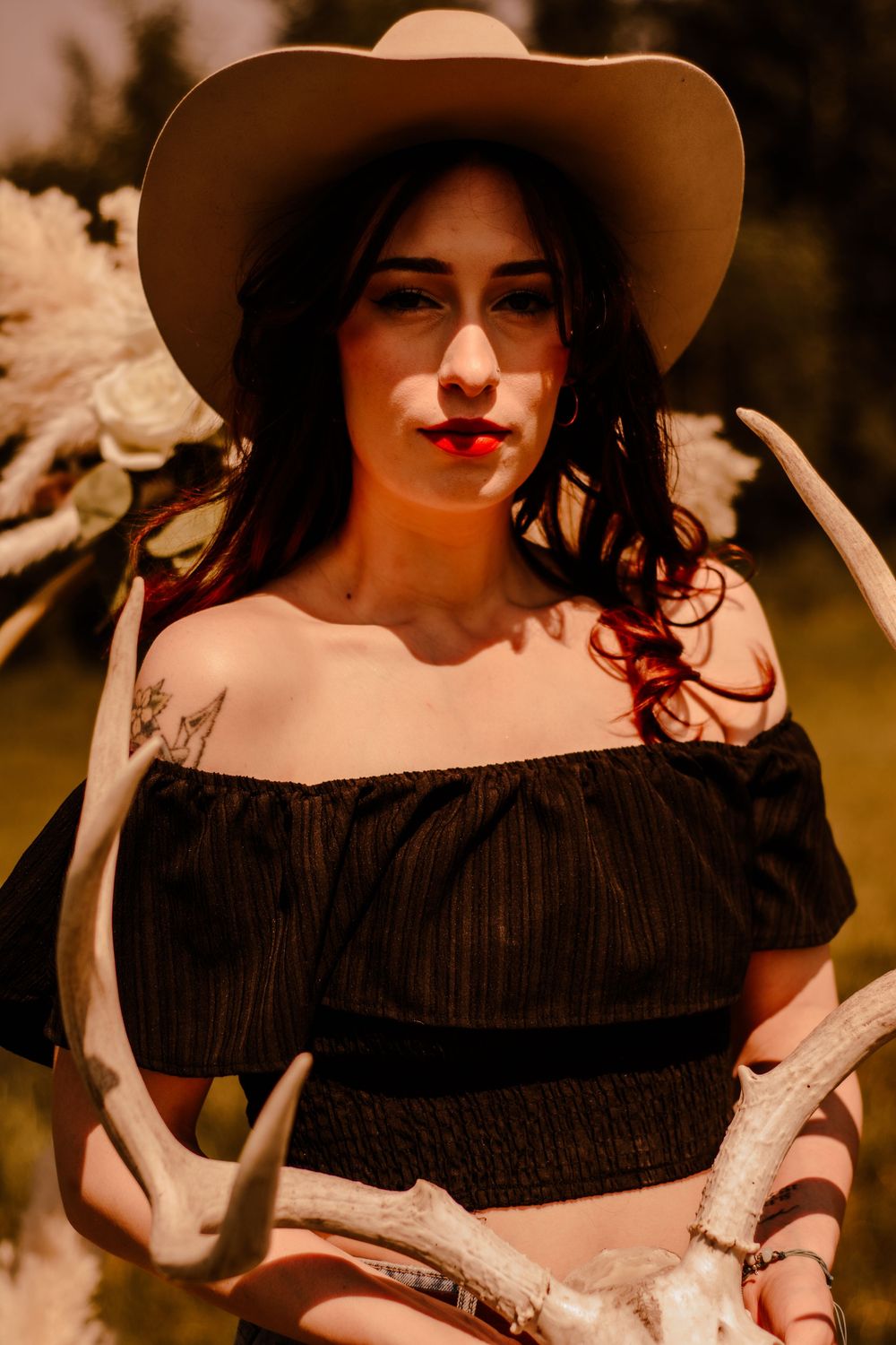 Person in a cowboy hat and off-shoulder black dress poses with antlers in warm sunset lighting.