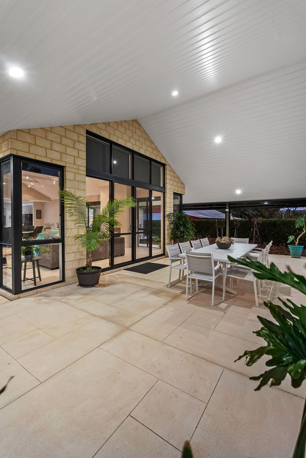 Modern outdoor patio area with glass doors, recessed lighting, cream tile flooring and white dining furniture at dusk.