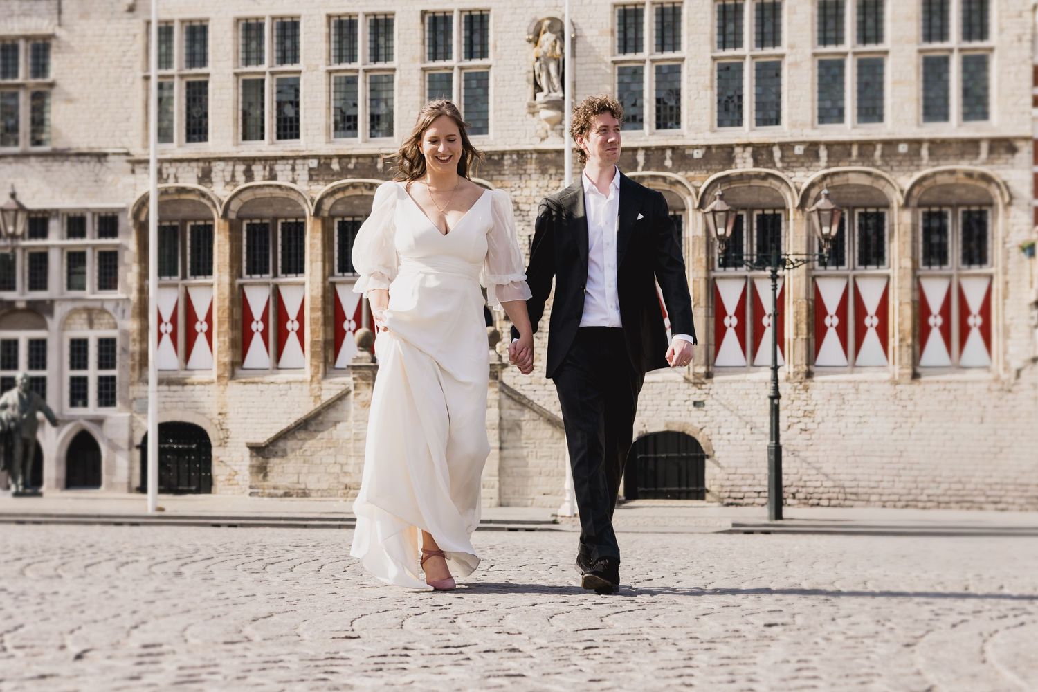 A couple in formal wedding attire walking together against a historic Gothic building with ornate architecture.