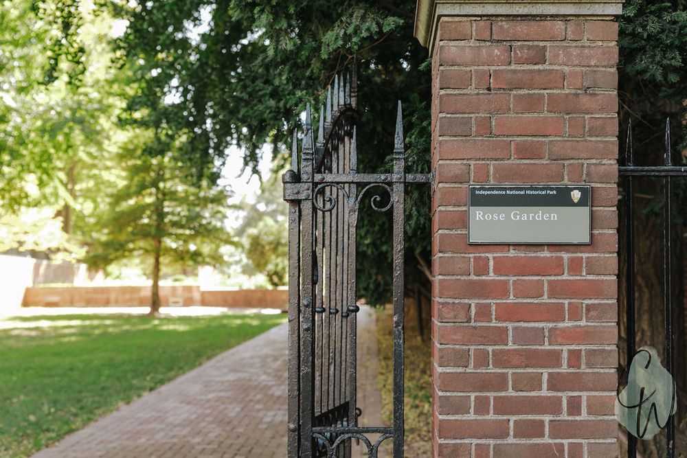 A wrought iron gate with a mail slot on a red brick pillar opens to a tree-lined walkway on a sunny day.