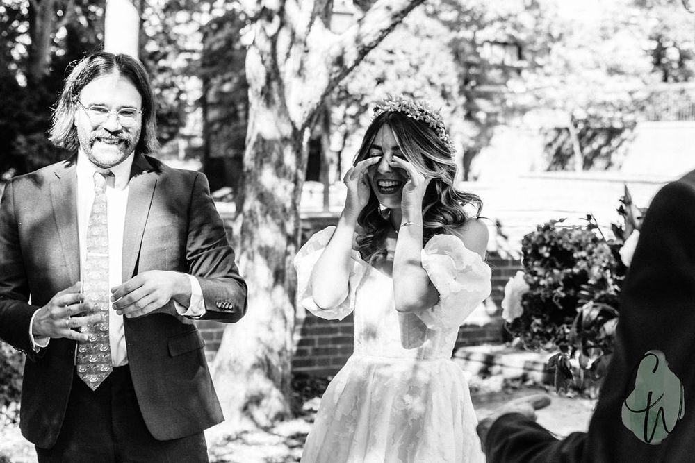 A candid black and white wedding photo showing a bride taking a photograph while another person laughs next to her.
