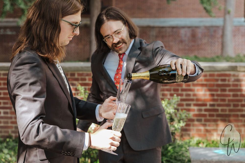 Two people in formalwear pop open a bottle of champagne outside a brick building on a sunny day.