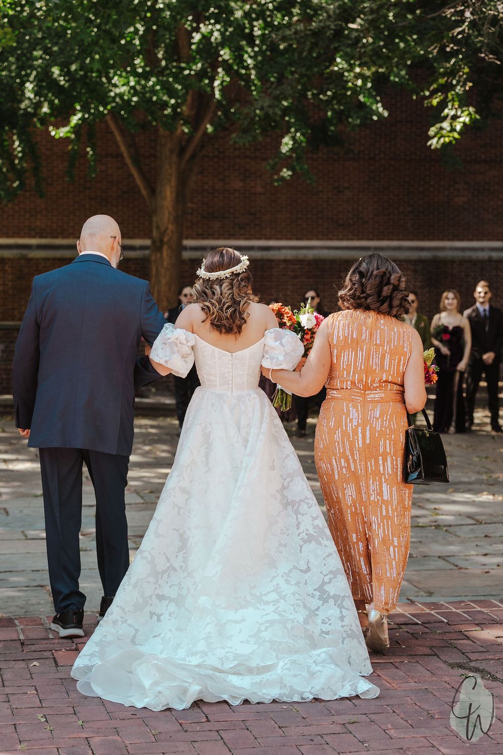 Wedding party walking down a brick path with bride in lace gown and guest in orange dress under trees.