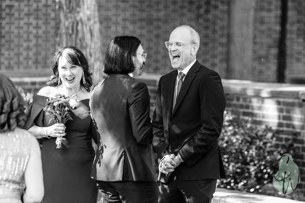 Three people share a candid black and white moment during an outdoor gathering with brick building in background.