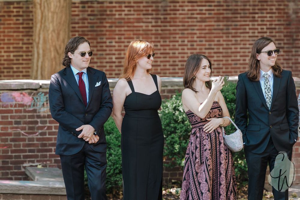 Four wedding guests in formal attire stand together against a brick wall at an outdoor venue.