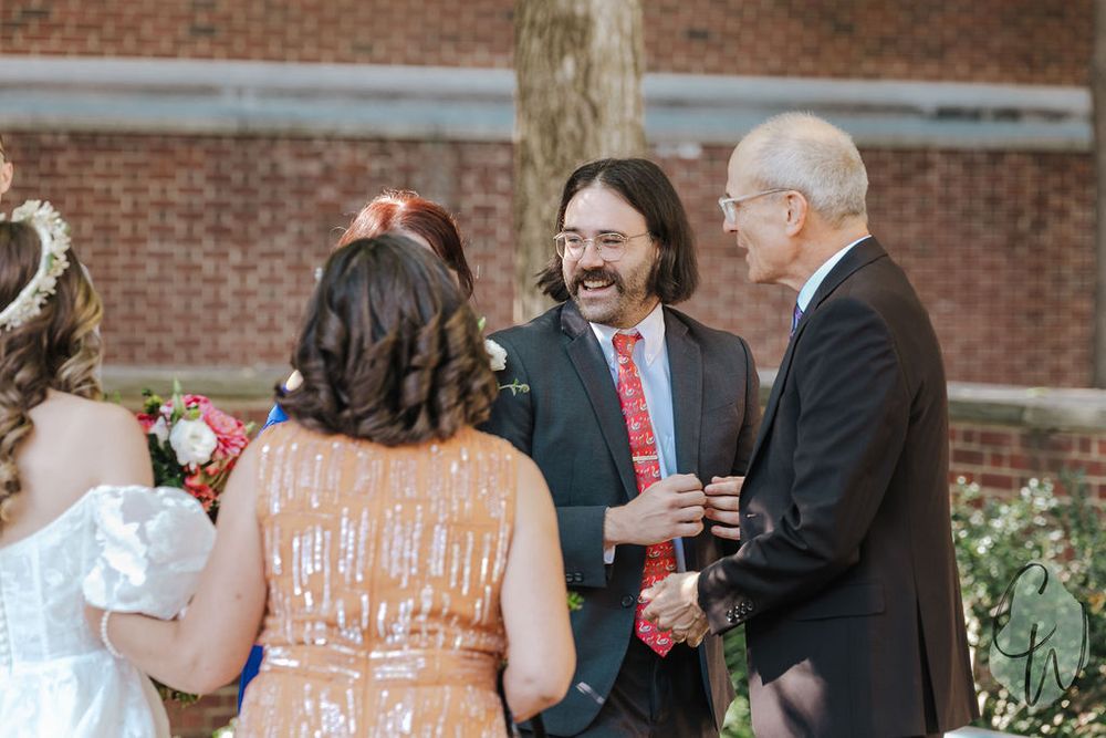 Wedding party members in matching coral dresses stand together in front of a brick building during an outdoor ceremony.