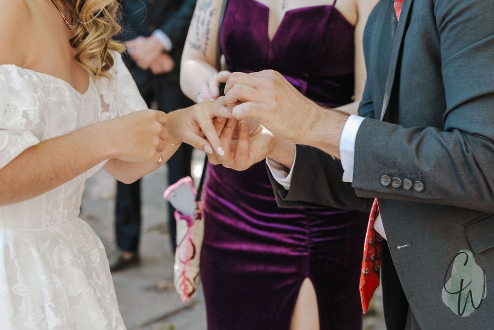 Two people exchanging wedding rings during a ceremony with a guest in a purple dress visible in the background.