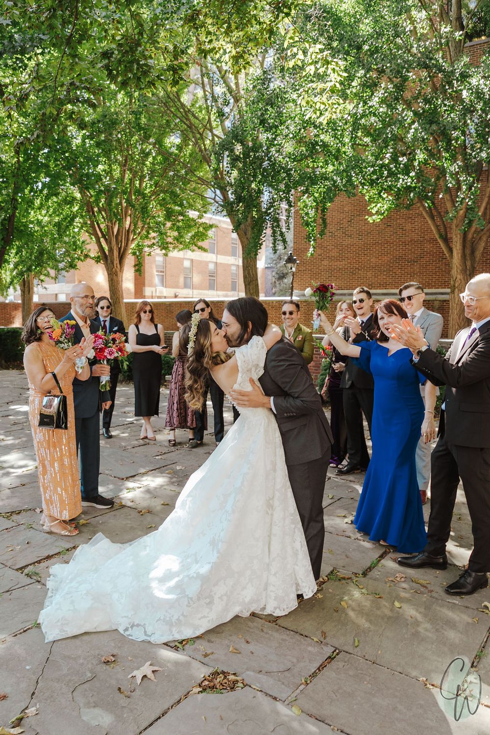 A wedding party gathers in a shaded outdoor courtyard area during a celebratory moment.