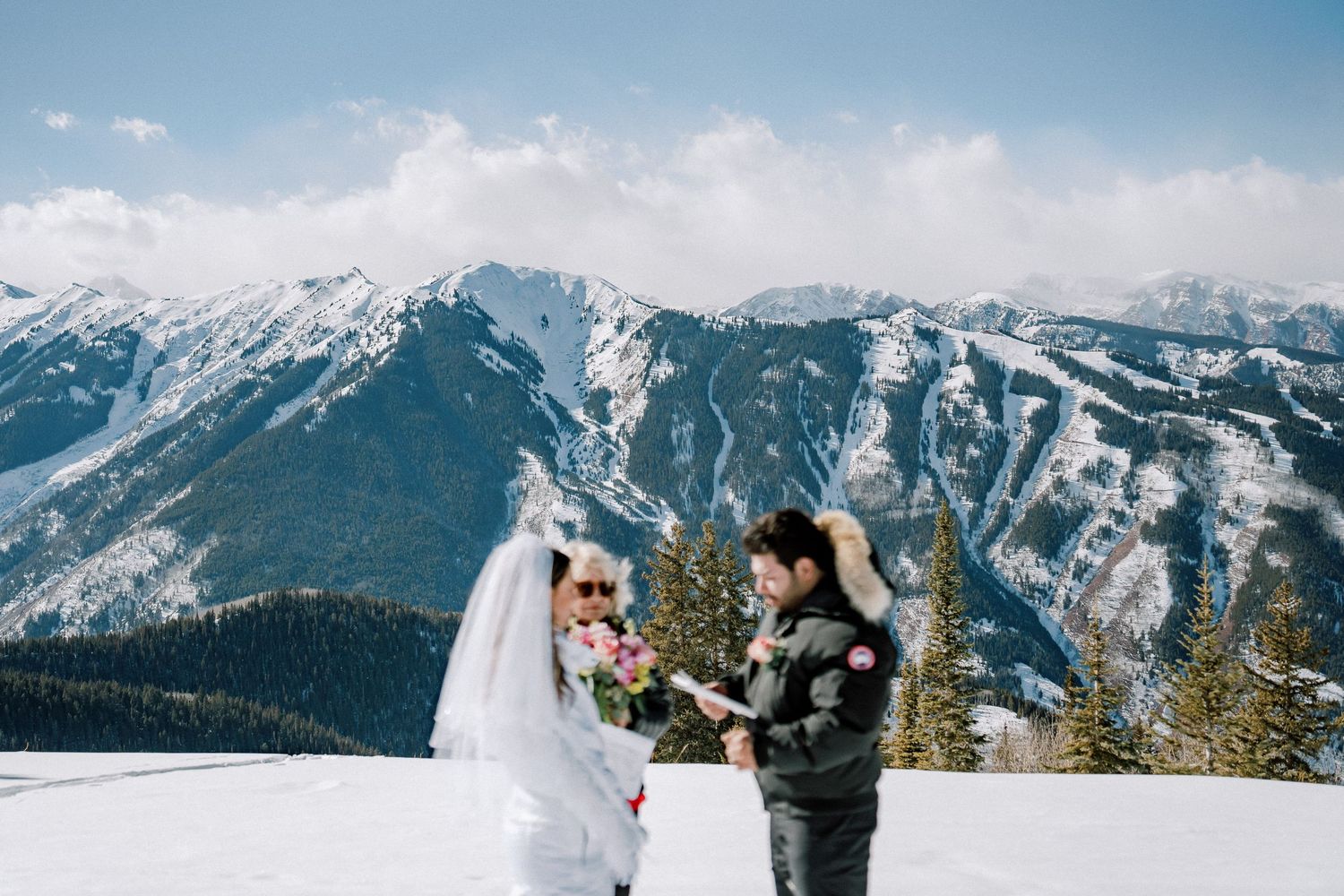 A wedding couple stands together on a snowy mountain summit with dramatic snow-capped peaks in the background.