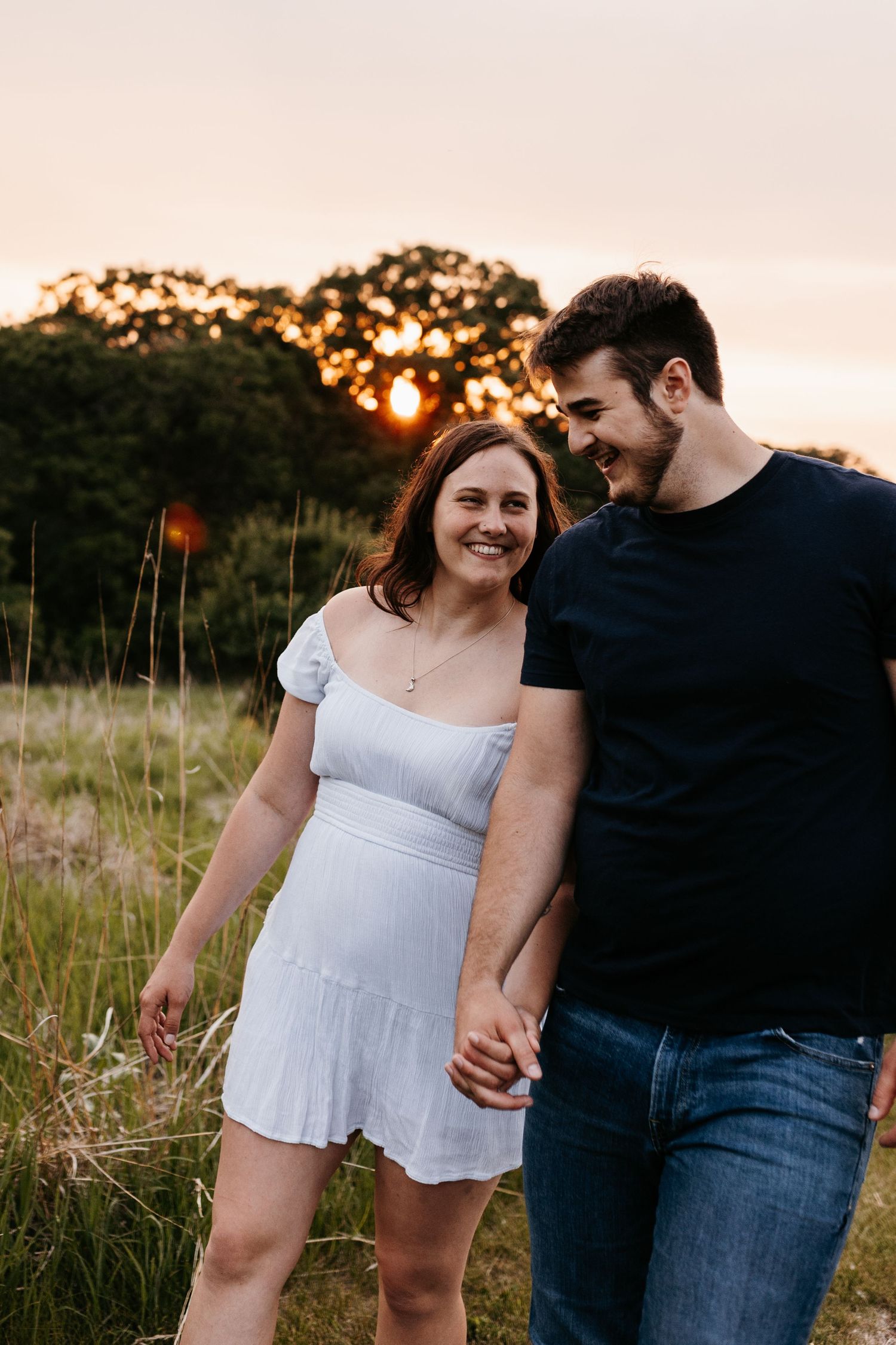 A couple holding hands and laughing while walking through tall grass at sunset in a field.