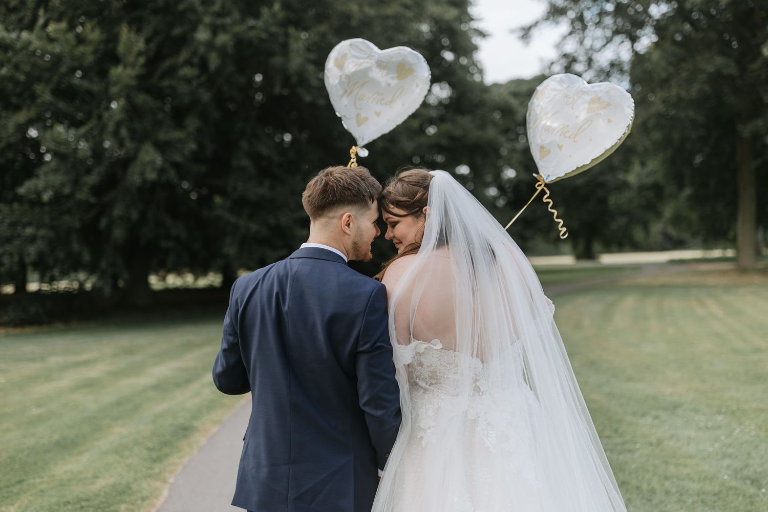 Newlyweds share an intimate moment on a grassy lawn with white heart-shaped balloons floating behind them.