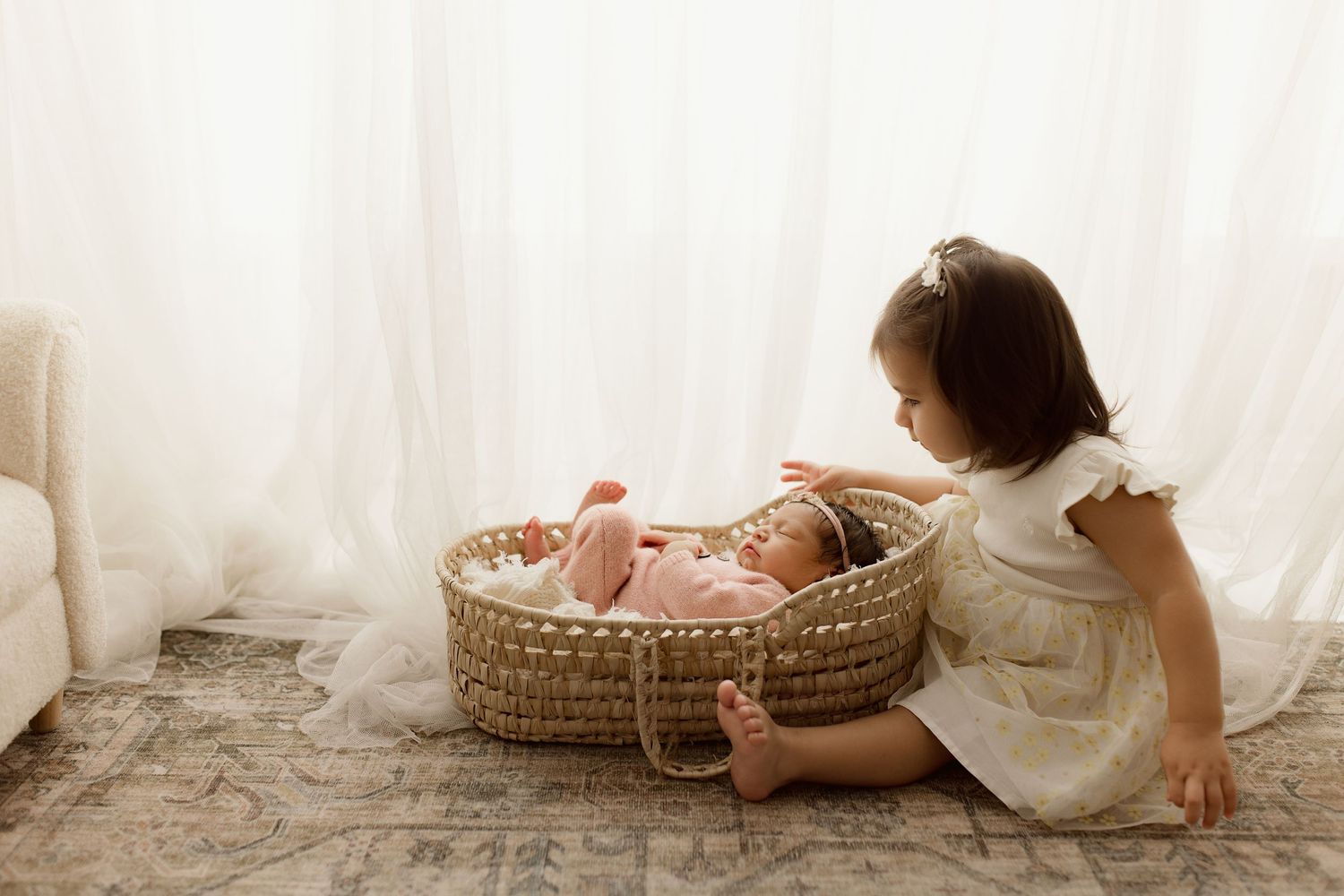 A young child in white dress sits beside a woven basket with baby sibling on a carpeted floor near sheer curtains.