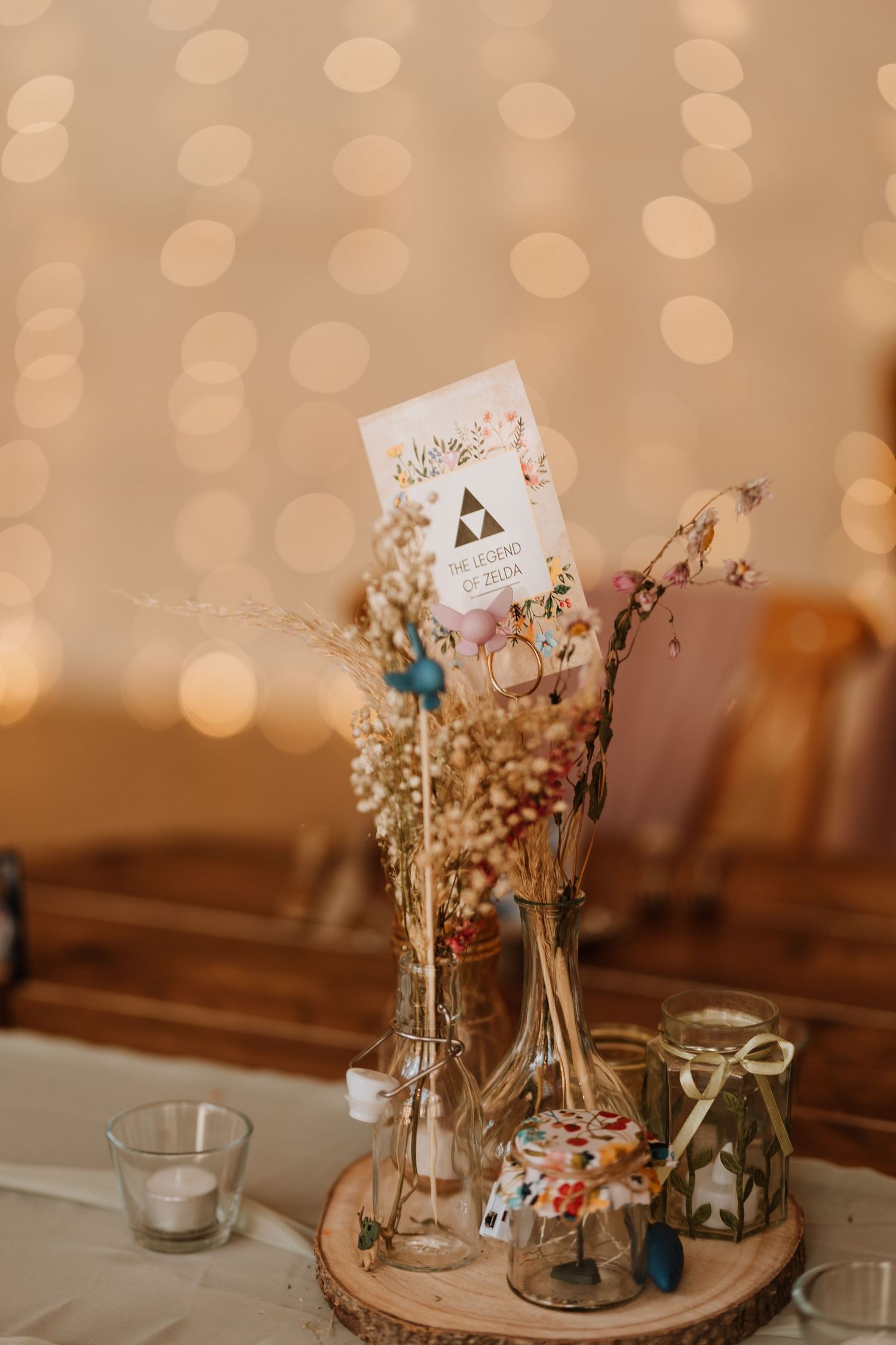 Rustic wedding table decor featuring dried flowers in glass vases and table number on a wood slice with bokeh background.