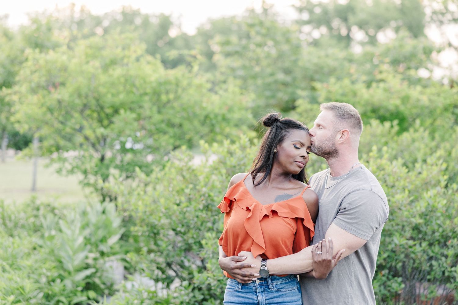 A couple embraces outdoors in a natural setting with lush green foliage in the background.