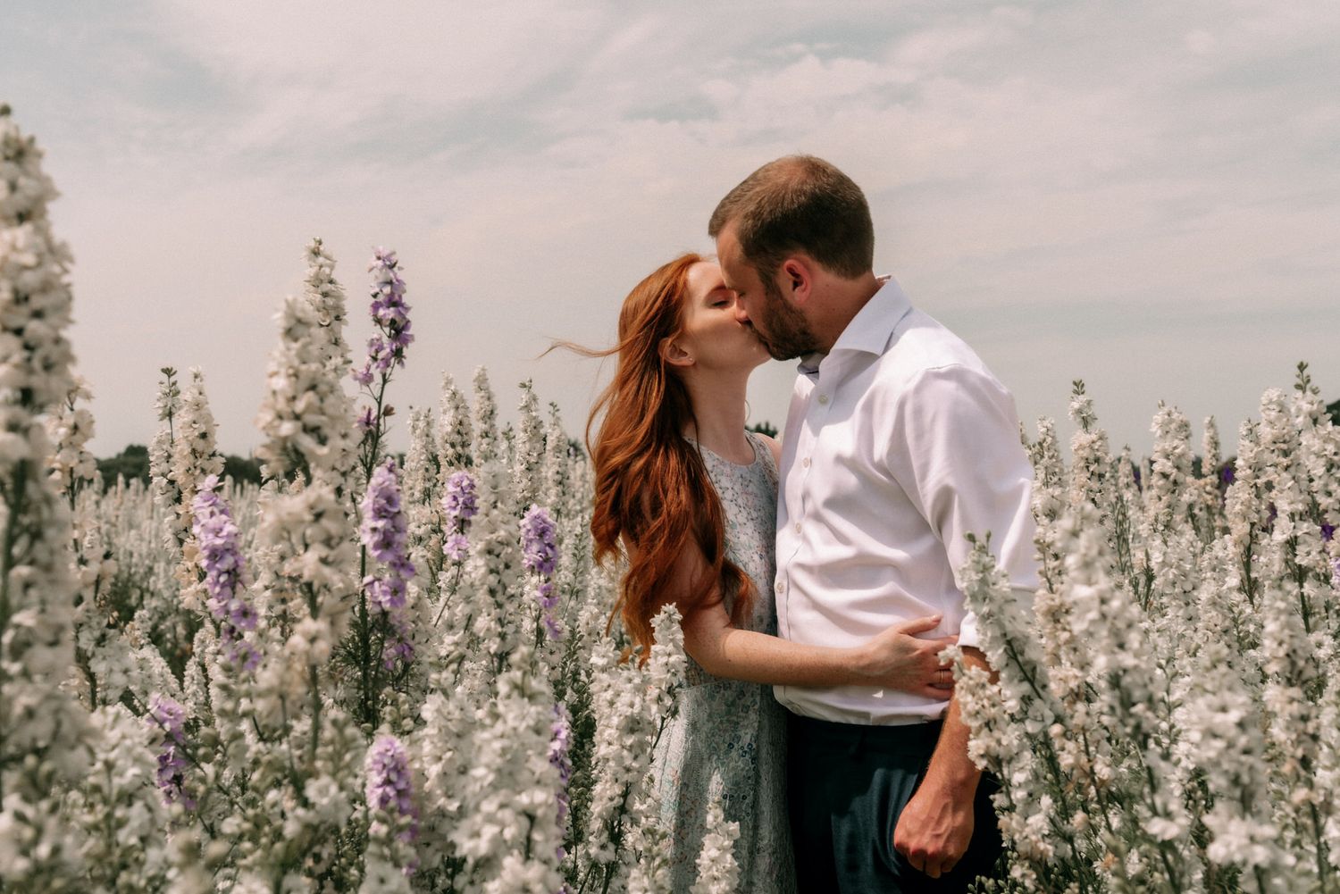 A romantic couple embraces in a blooming field of purple and white delphinium flowers on an overcast day.