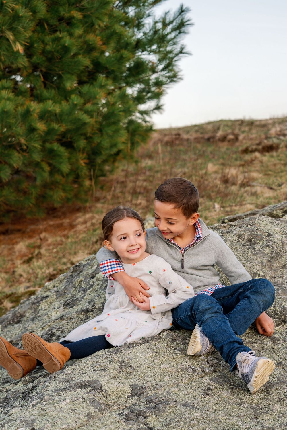 Two children sitting together on a large rock formation with a pine tree in the background.