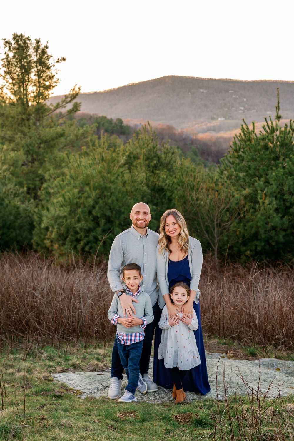 Family portrait of four standing together outdoors with mountains and trees in background.
