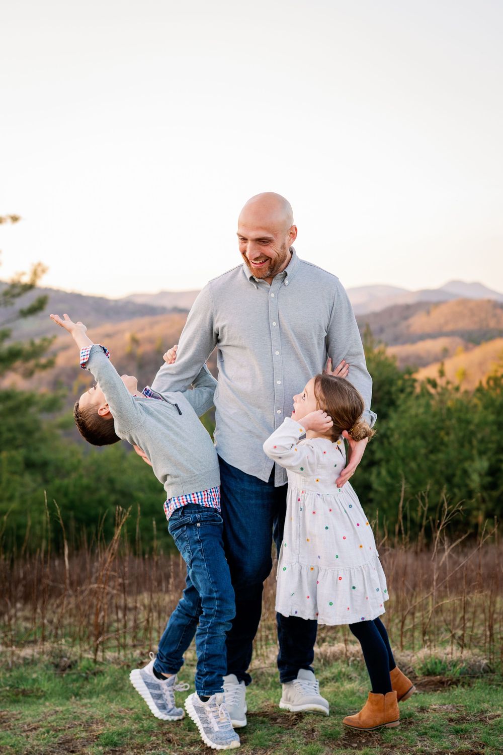 Parent playing and laughing with two children on a hilltop during sunset.