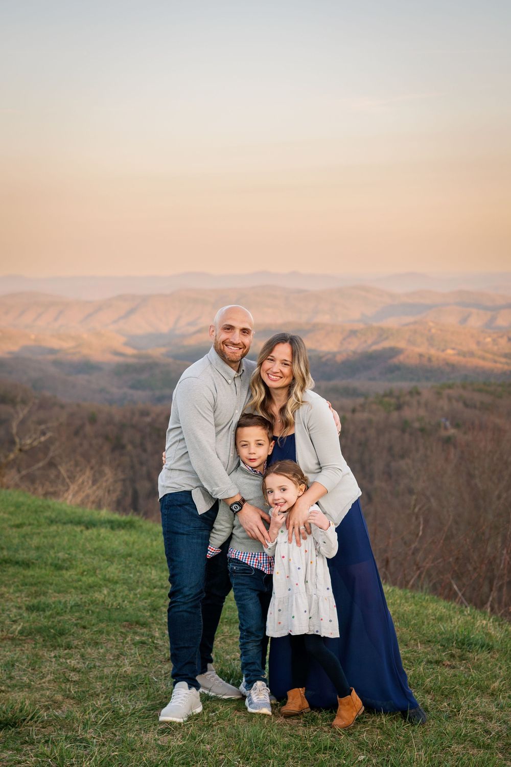 Family of four embracing while posing for a portrait against a mountain landscape at sunset.