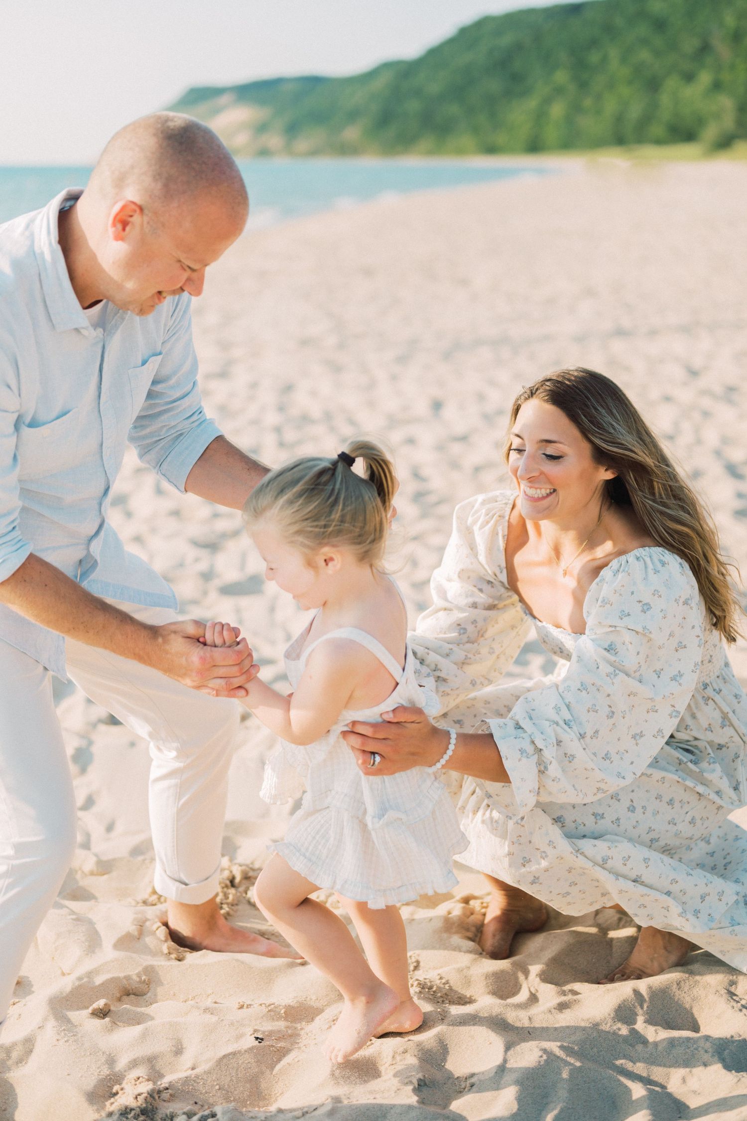 A family enjoys a tender moment together while playing on a sandy beach with hills in the background on a sunny day.