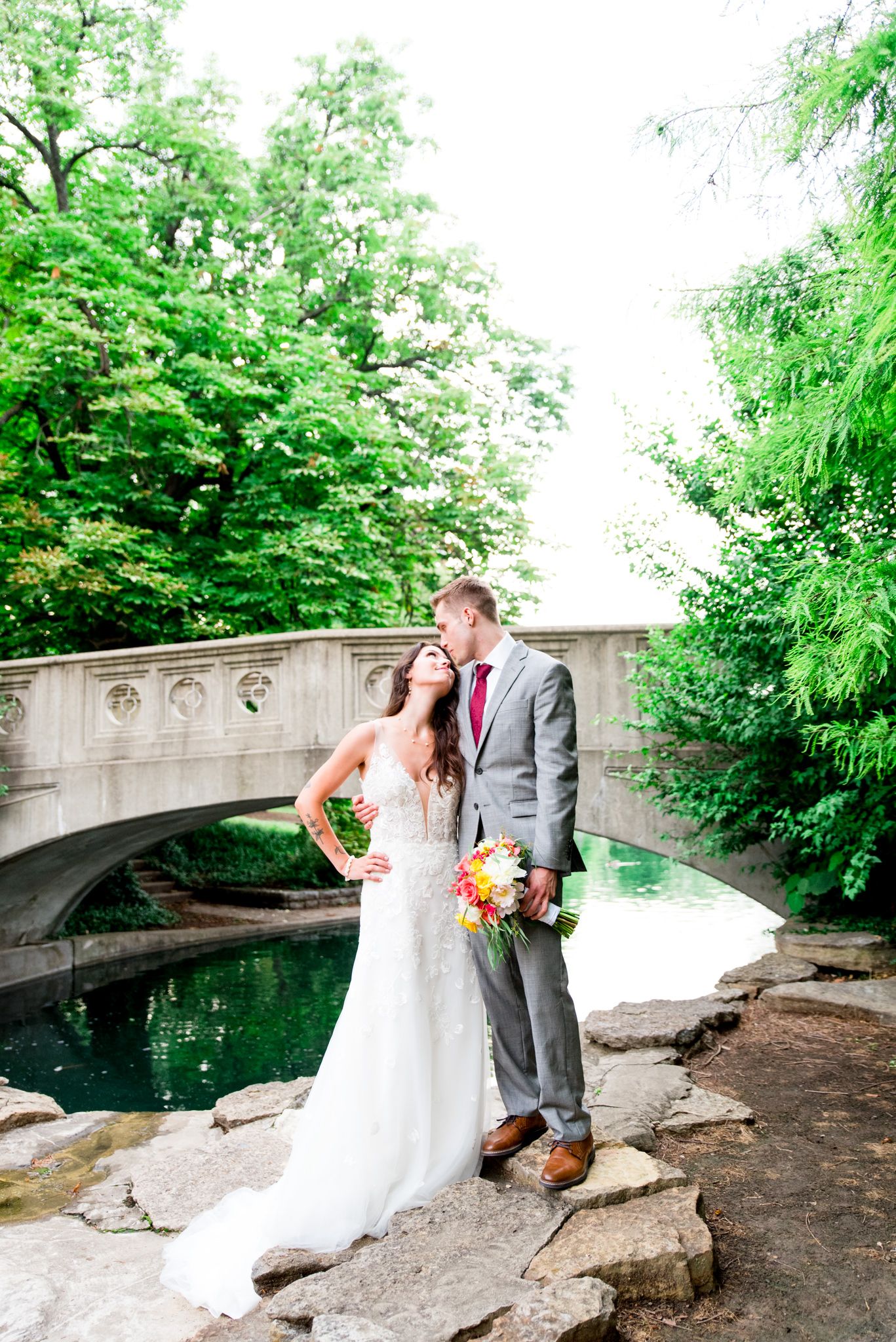 bride in Serafine gown and groom in gray suit holding pink and yellow bouquet looking at each other at Eden Park