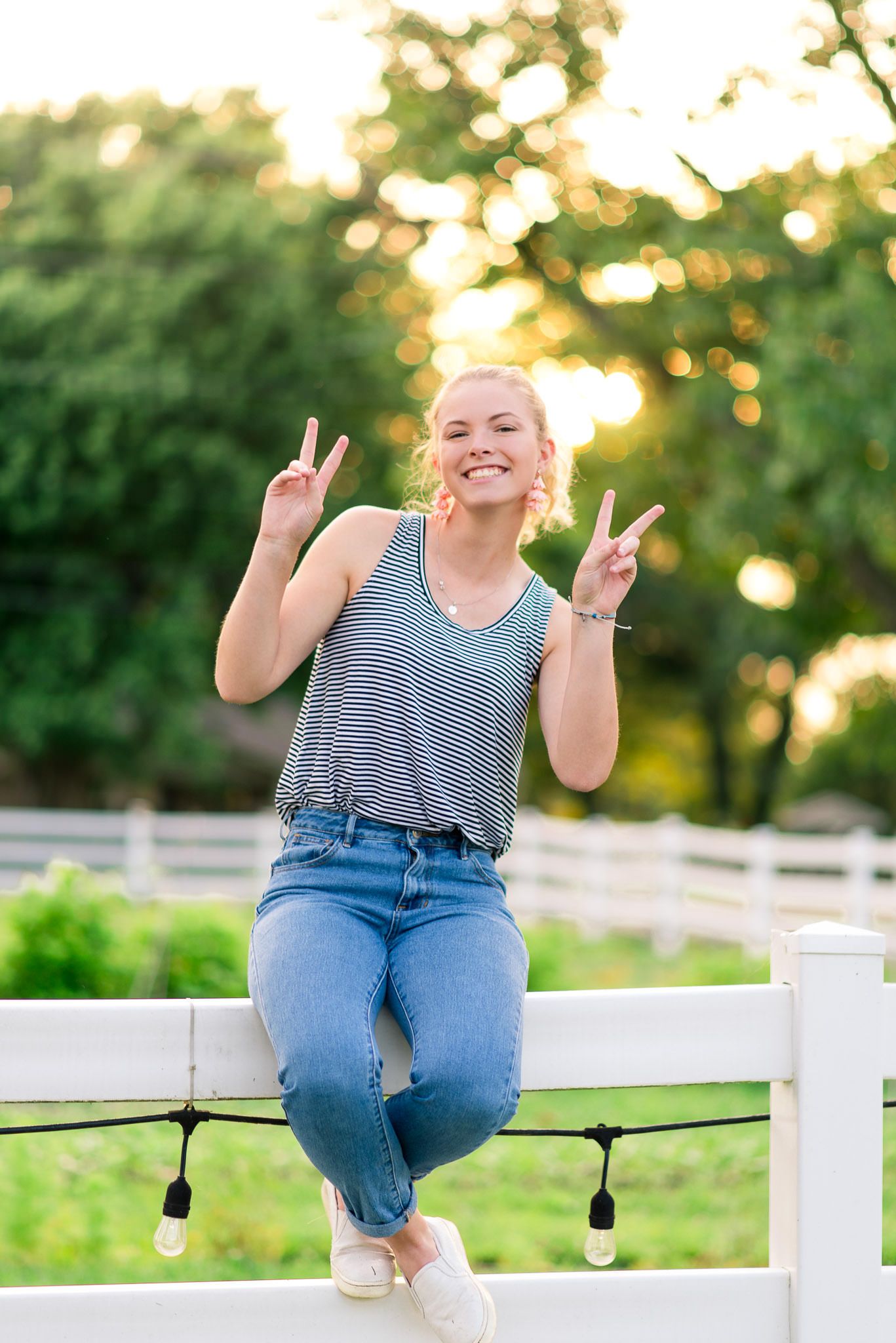 young woman sitting on a white fence holding up two peace signs in front of trees at sunset