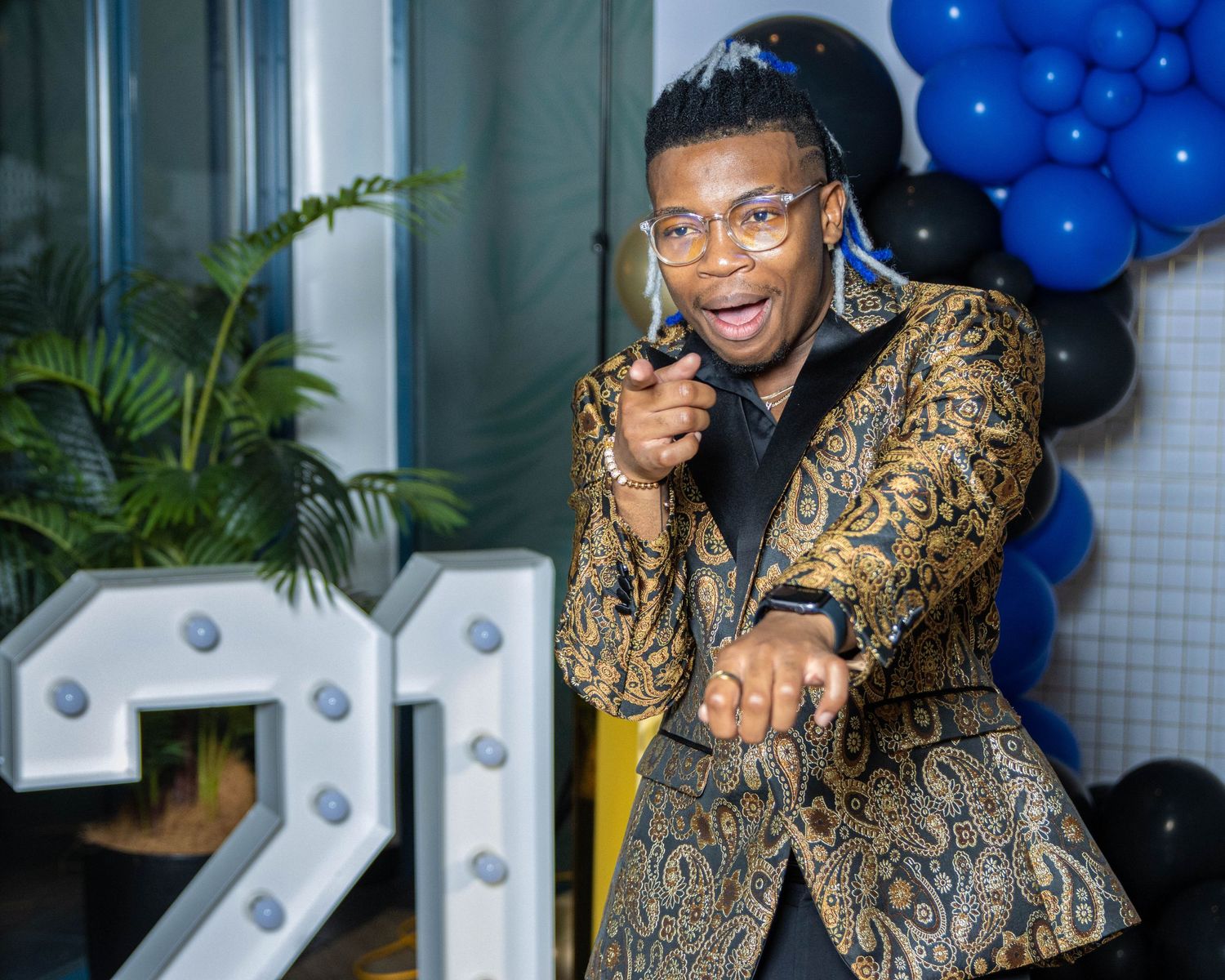 Person in patterned blazer strikes dynamic pose against backdrop with blue balloons.
