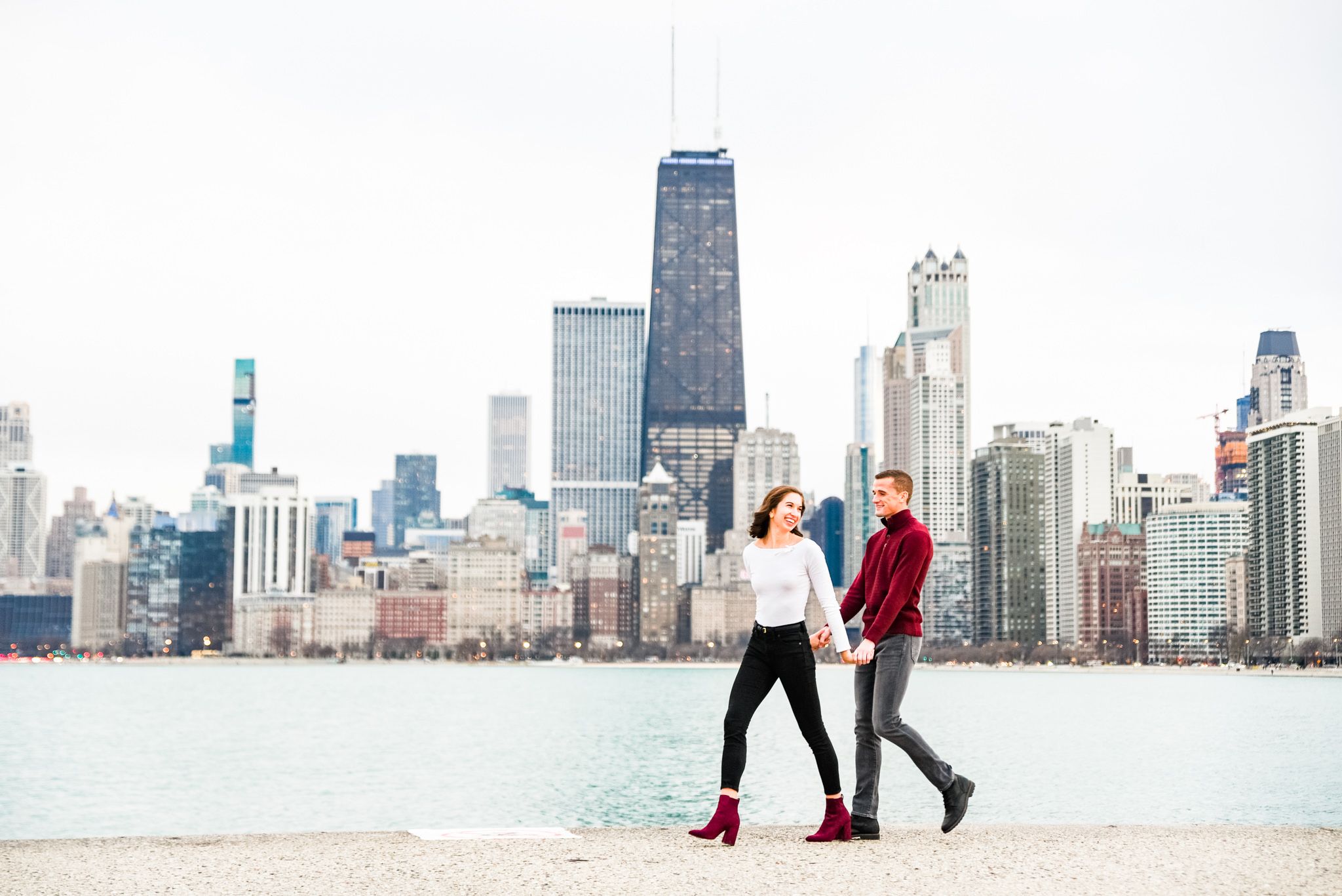 woman in white shirt and black jeans holding hands leading man along pier at North Avenue Beach for Chicago engagement
