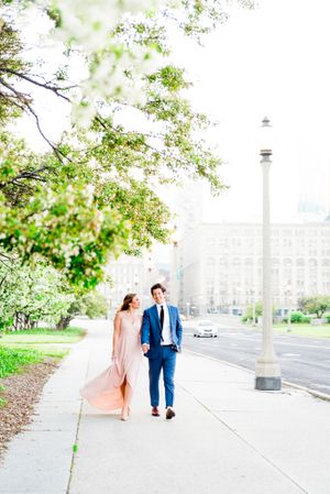 woman in pink dress looks at fiancee in navy suit as they walk along sidewalk near Millennium Park for sunset engagement
