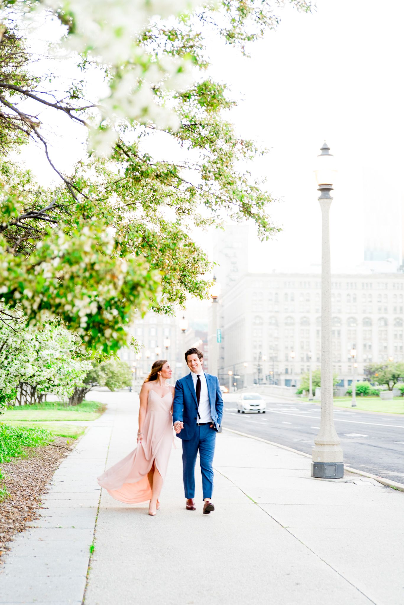 woman in pink dress looks at fiancee in navy suit as they walk along sidewalk near Millennium Park for sunset engagement
