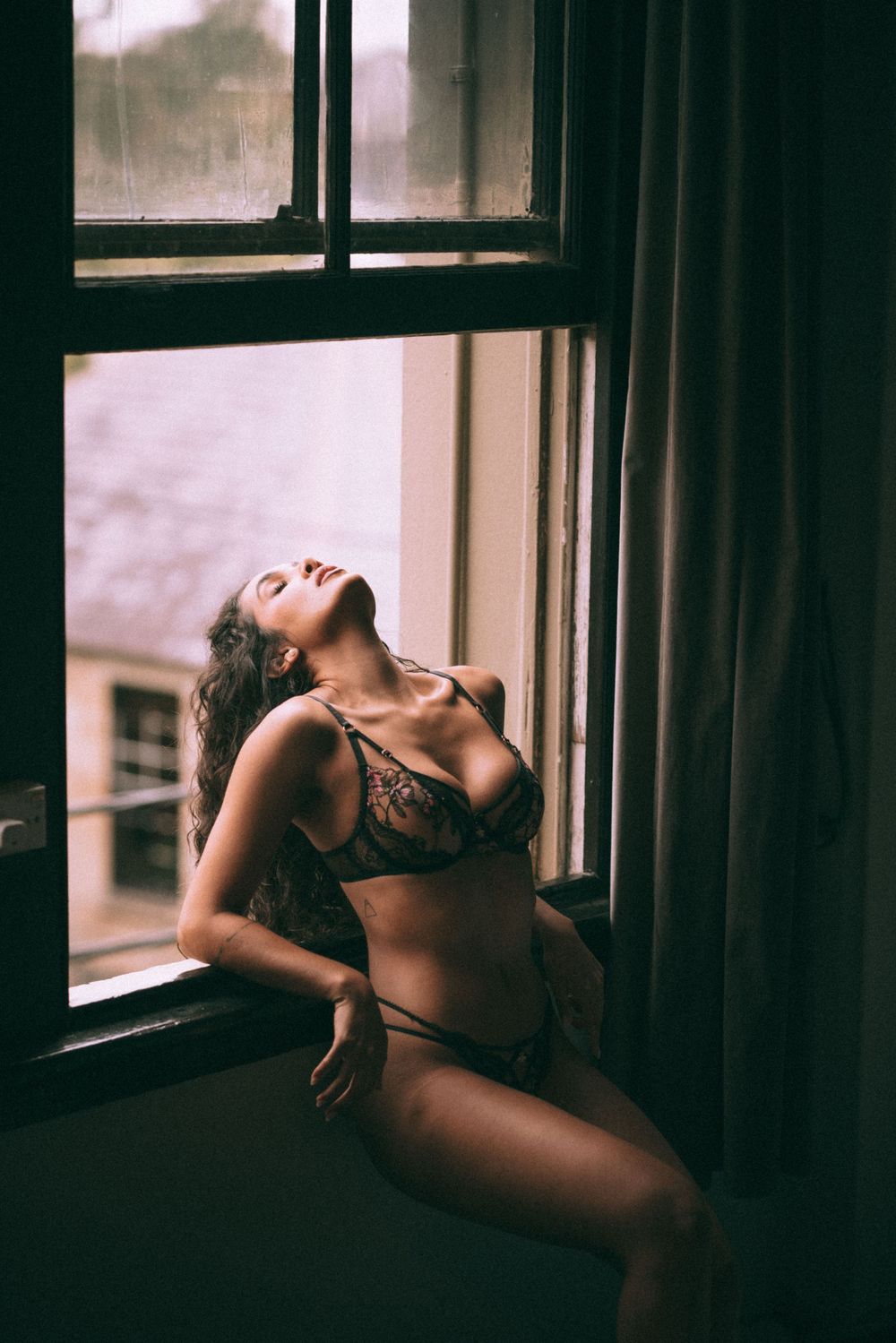 Artistic boudoir portrait by window with dramatic natural lighting and lace lingerie.