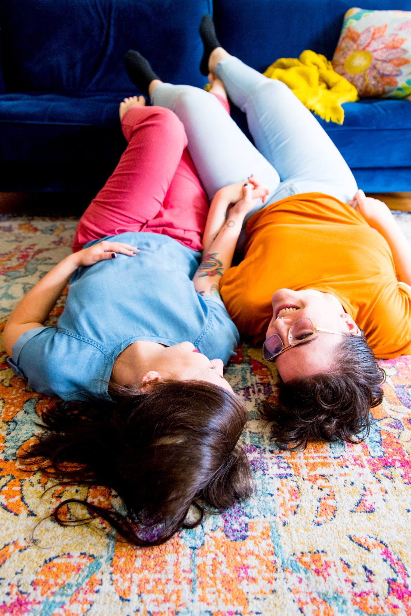 lesbian couple laying on orange and pink rug with feet up on blue couch, smiling at each other