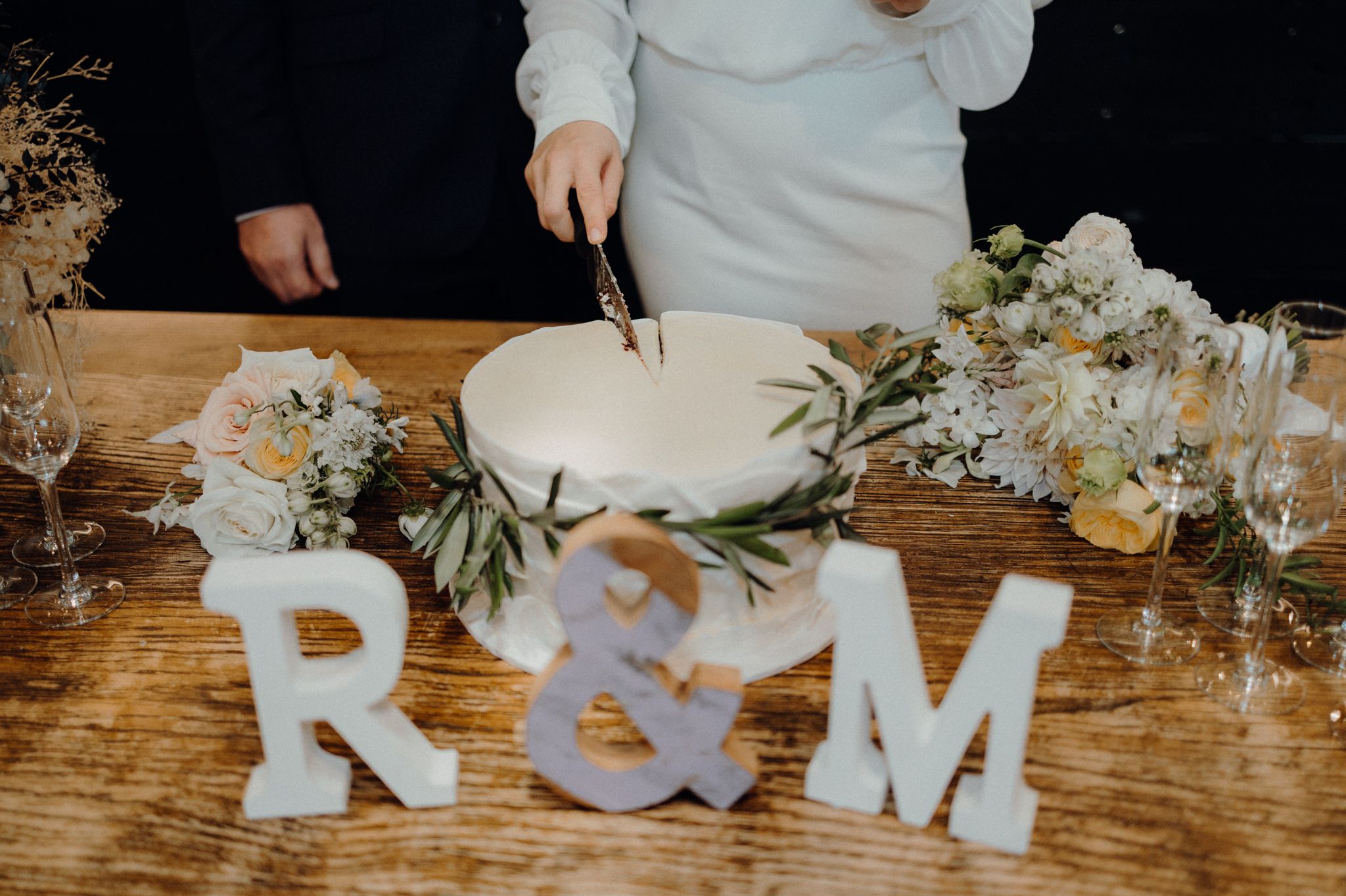Wedding cake cutting scene with white R&M letters and floral decorations on wooden table.