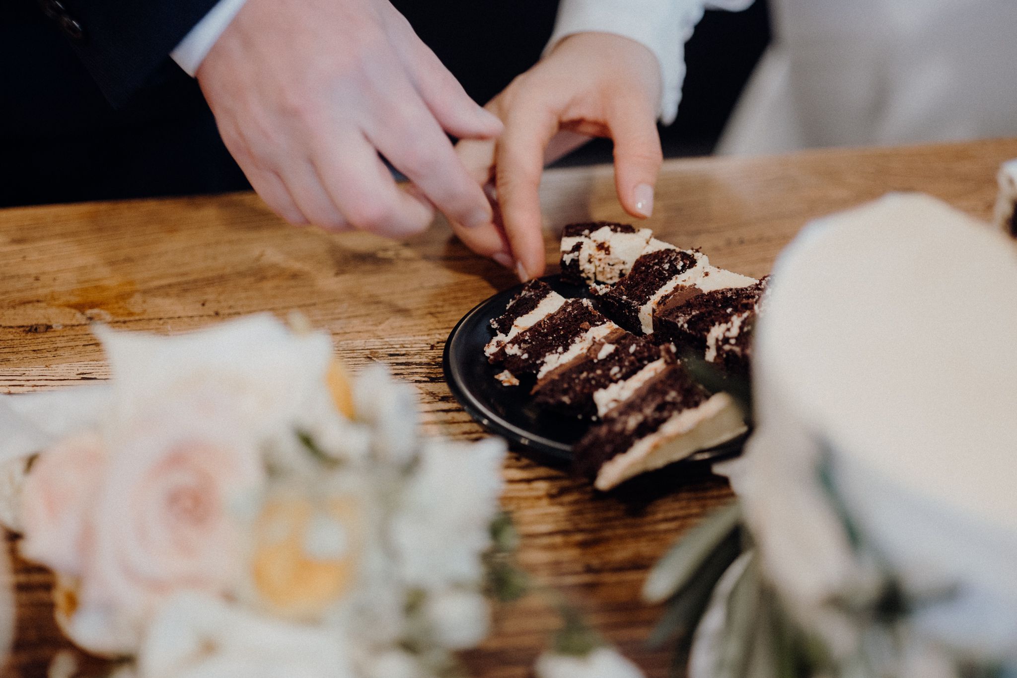 Close-up of hands reaching for chocolate desserts on dark plate with wedding flowers in background.