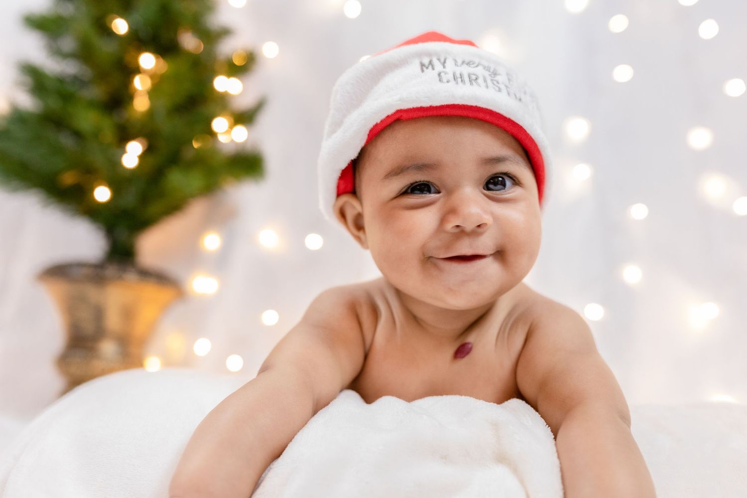Baby wearing red Santa hat lying on white blanket with Christmas tree in background.