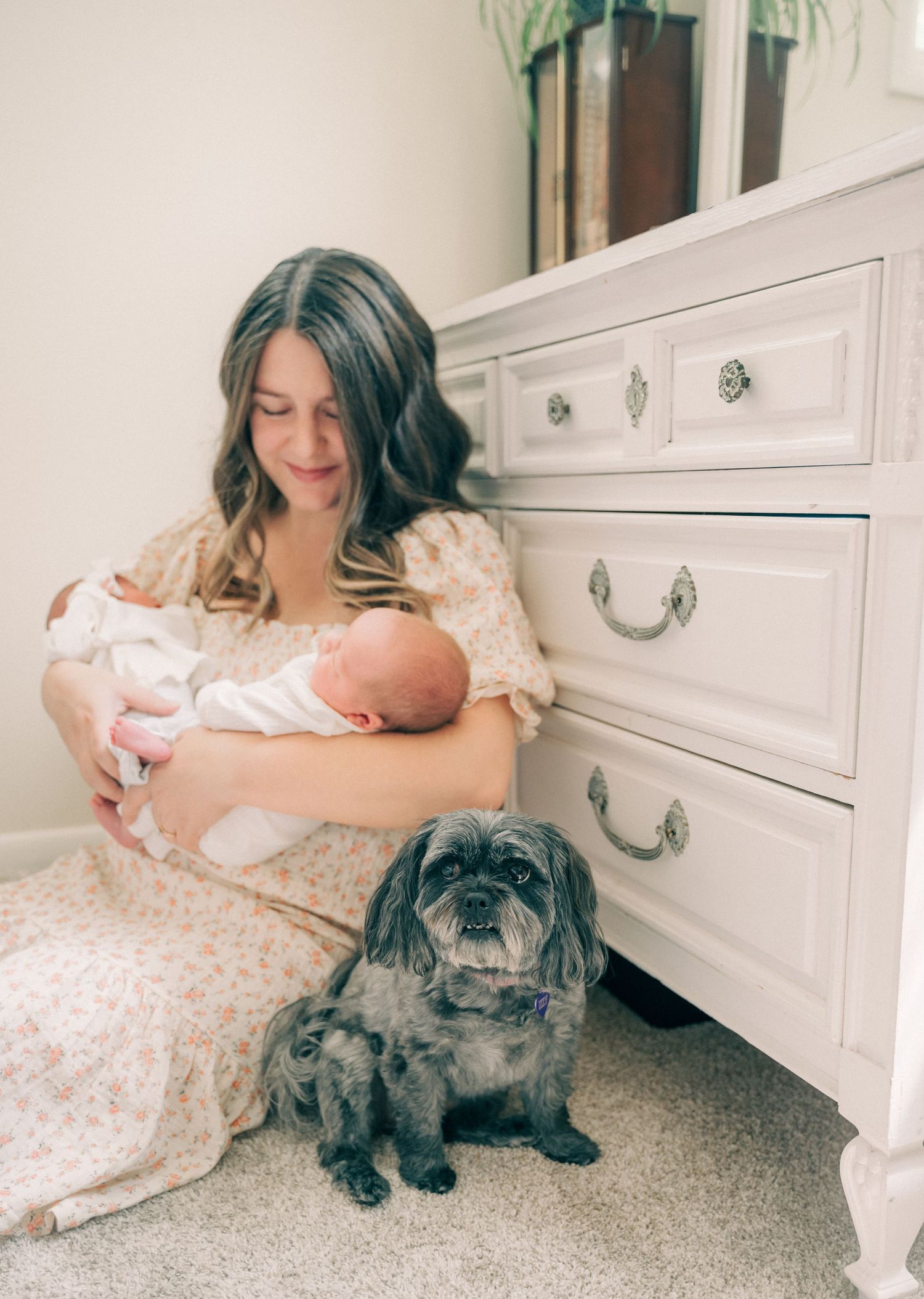A tender moment as a woman in a floral dress holds a baby while sitting beside a small black dog.