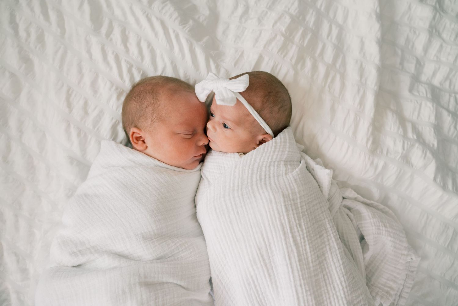 Two swaddled newborn babies lying close together on white bedding, one wearing a delicate bow.