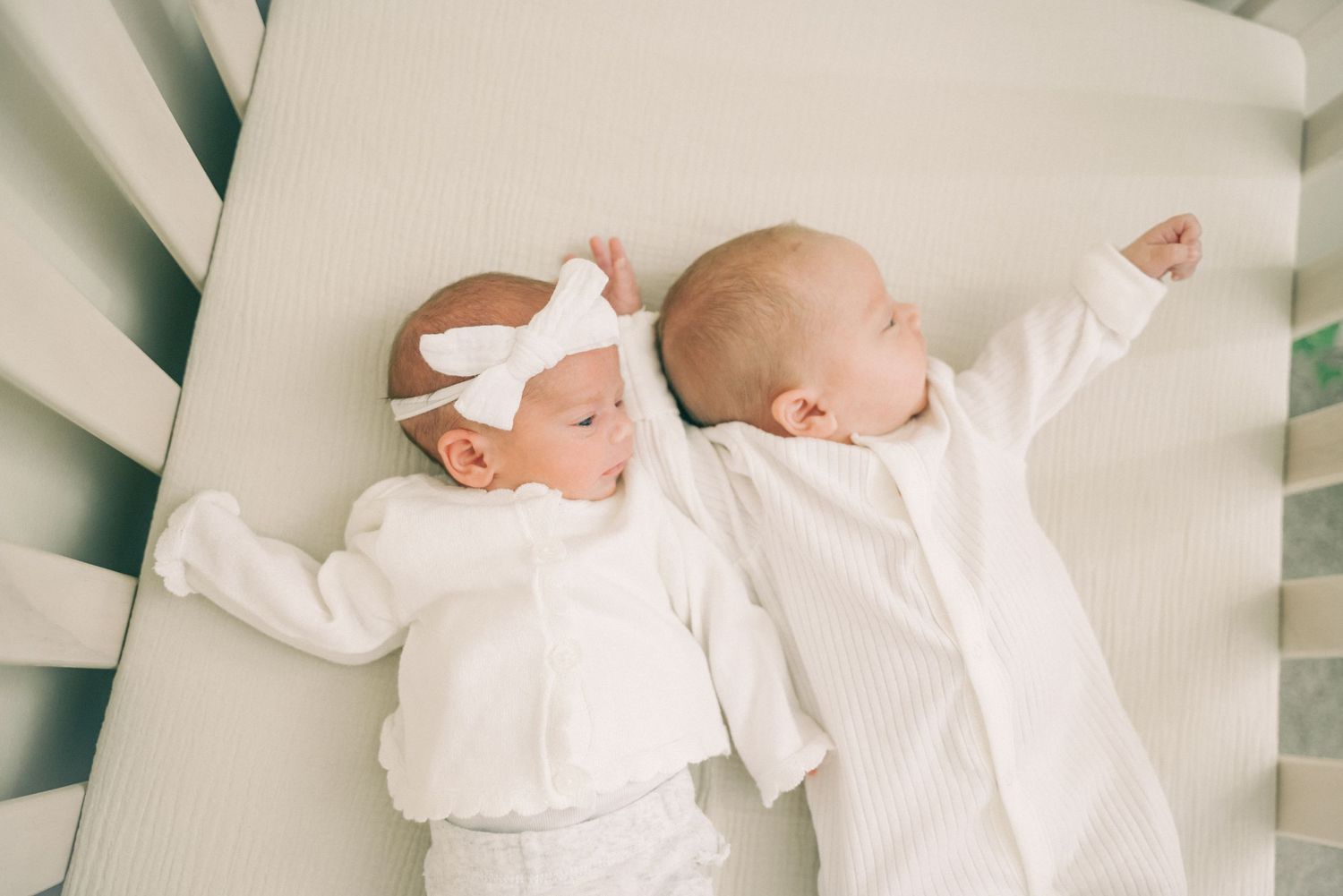 Two newborn babies dressed in white outfits lying side by side on white bedding in a crib.