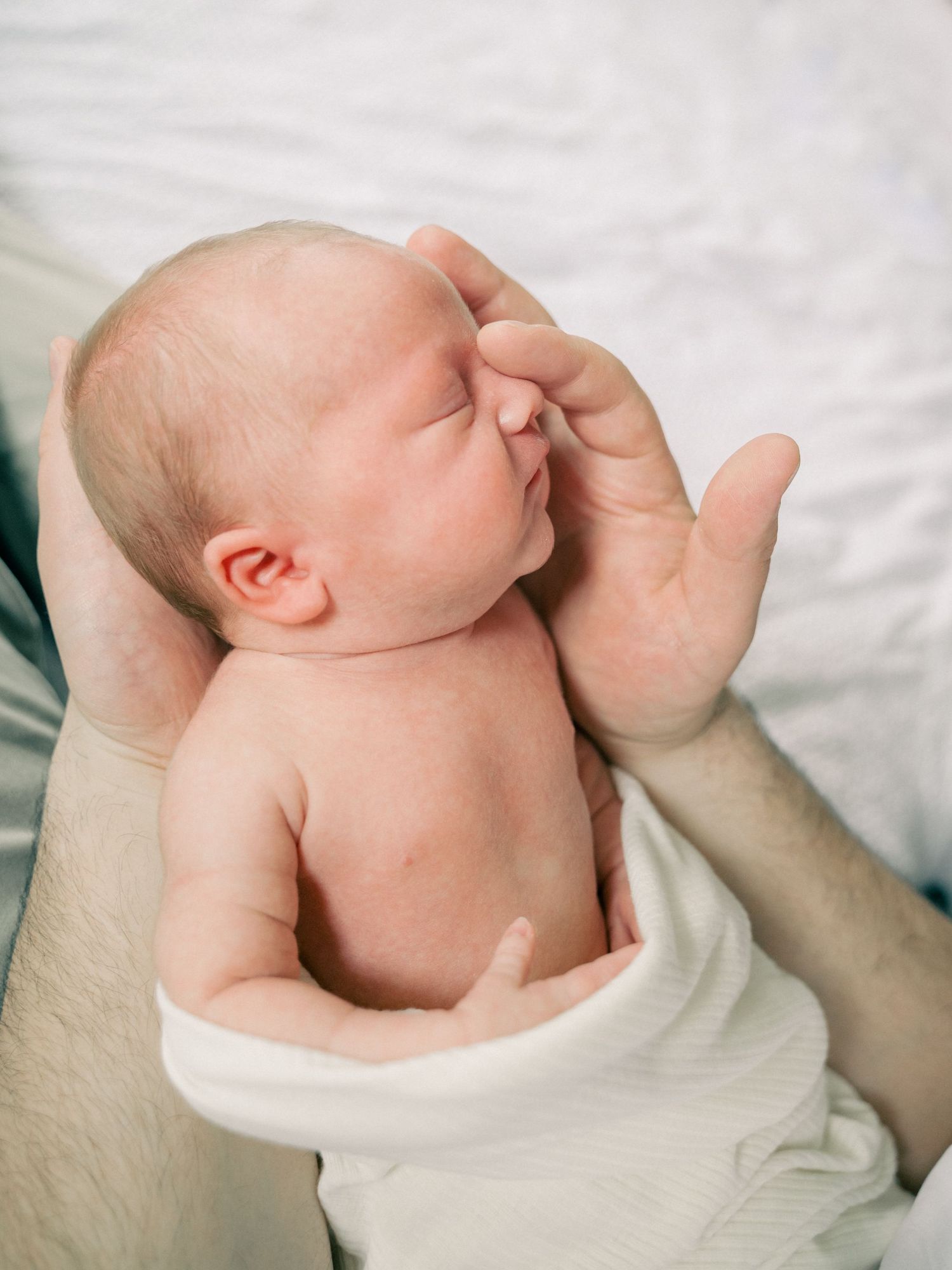 A newborn baby resting peacefully with hand near face on soft white fabric.