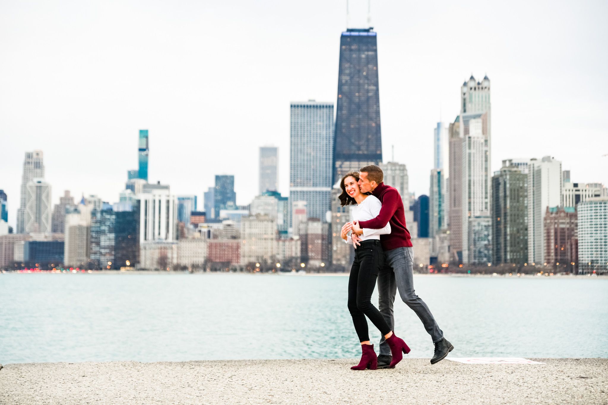 man in red sweater hugging woman from behind in front of Chicago skyline at North Avenue Beach for engagement pictures