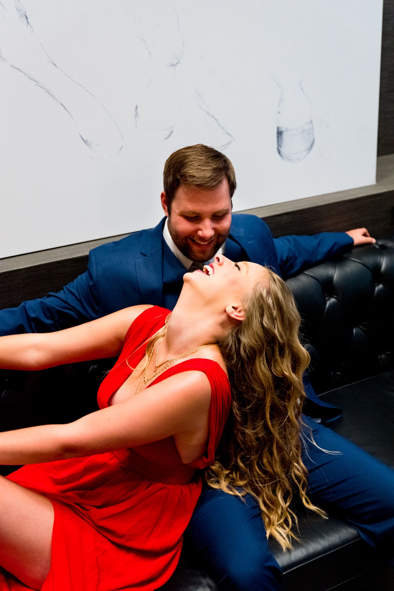 man in navy suit sitting on black leather couch and woman in red dress leans back and laughs