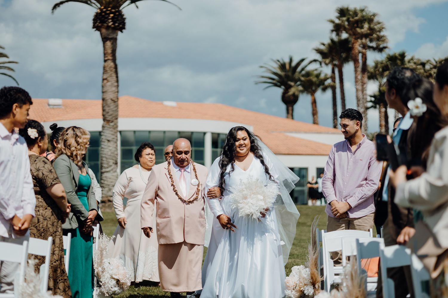 Outdoor wedding processional with guests in white chairs and palm trees in background.