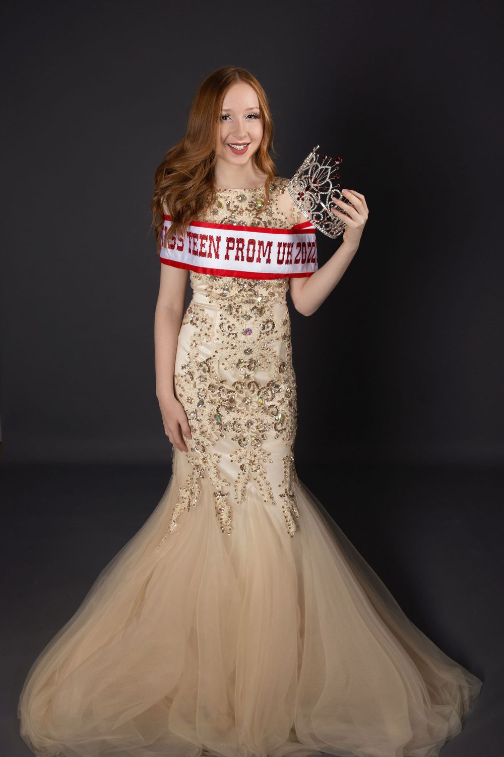 Teen pageant queen in gold dress with sash wrapped around her holding crown