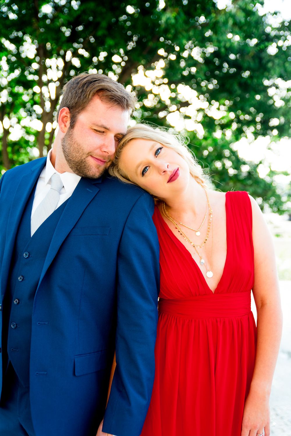 blonde woman in red dress and man in navy suit lean on each other in front of big tree