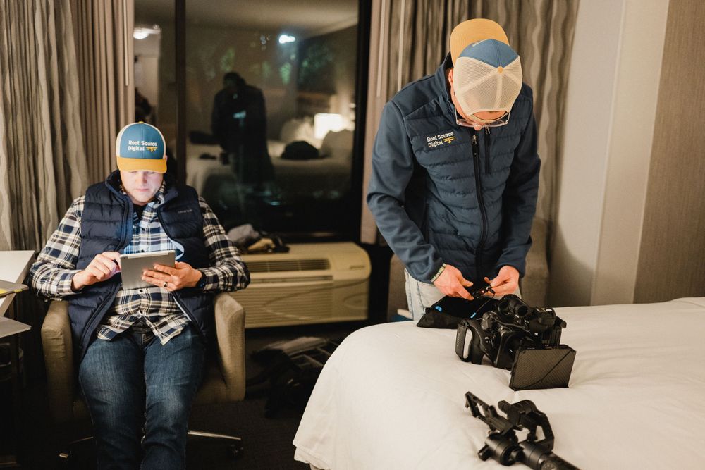 Two people preparing camera equipment on hotel room bed.