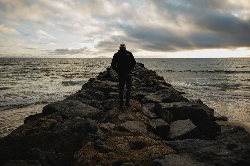 Silhouetted figure standing on a rocky jetty overlooking stormy ocean waters at sunset.