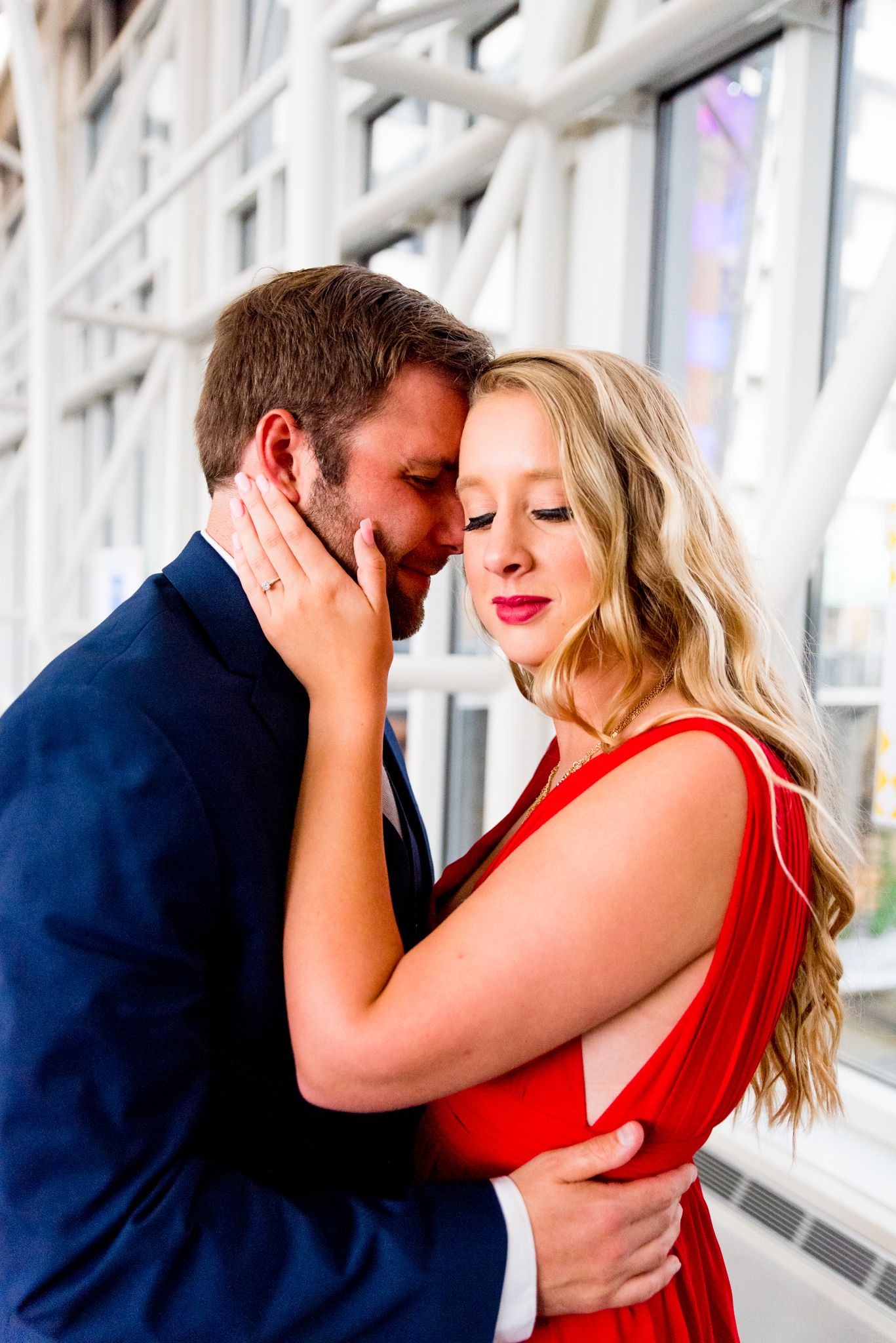 blonde woman in red dress looking at camera while fiance in navy suit rests on her forehead