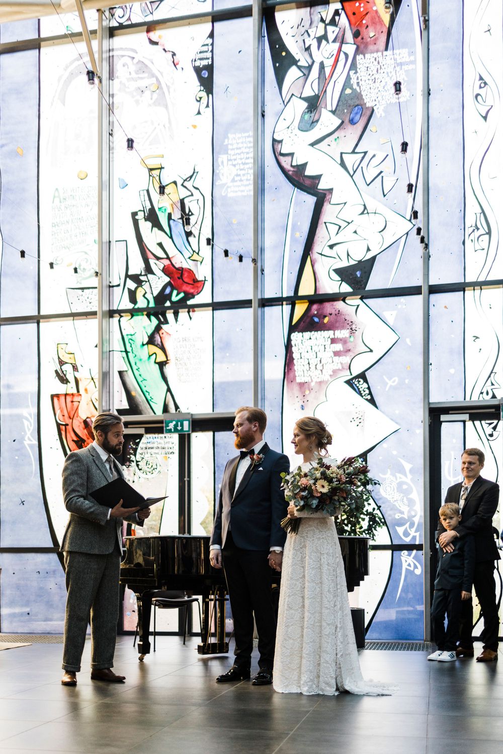 A wedding ceremony takes place in a modern venue with dramatic stained glass windows and a piano.