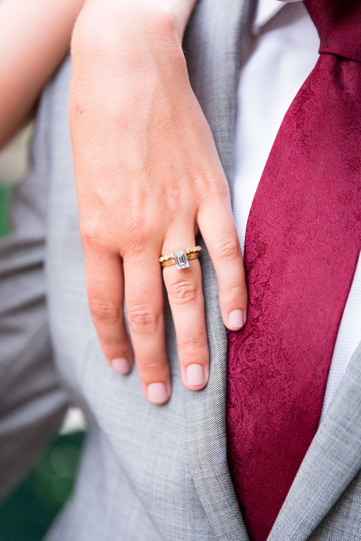 bride resting hand on grooms chest, showing her emerald cut engagement ring and his gray suit and red tie