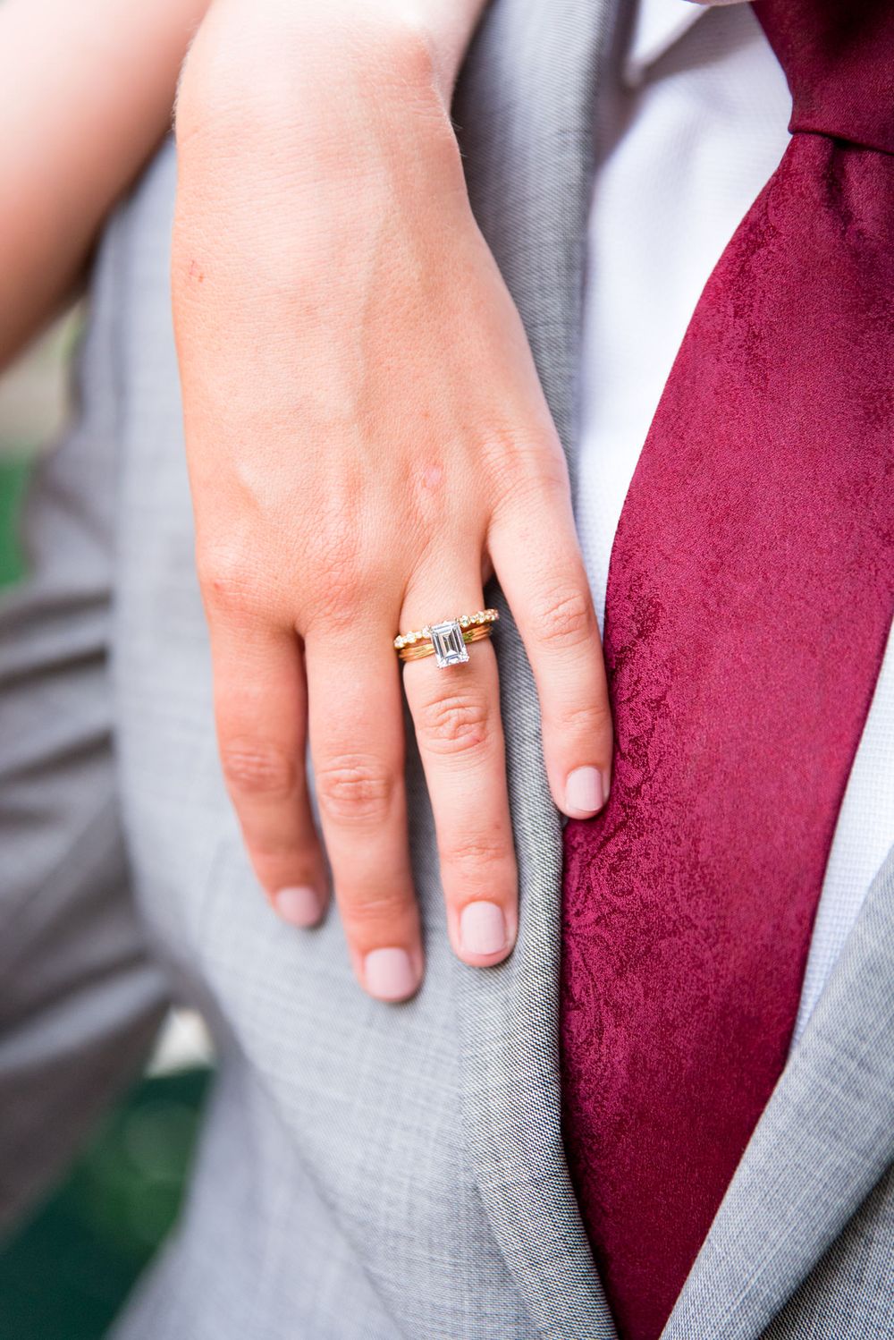 bride resting hand on grooms chest, showing her emerald cut engagement ring and his gray suit and red tie